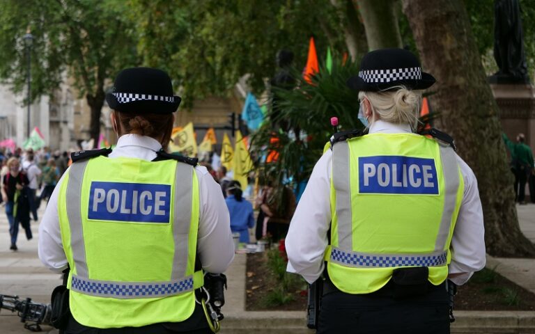 Two female police officers stood with their backs to the camera, both wearing hi-vis fluorescent jackets with the word 'police' on them.