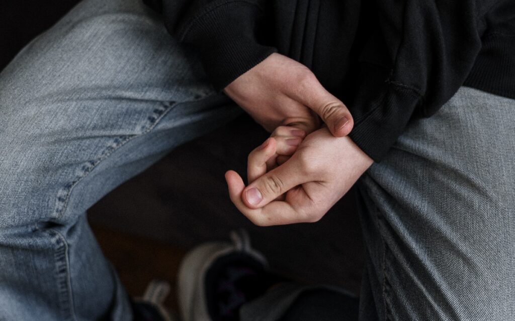 Person in black jacket and grey denim jeans, holding their hands together