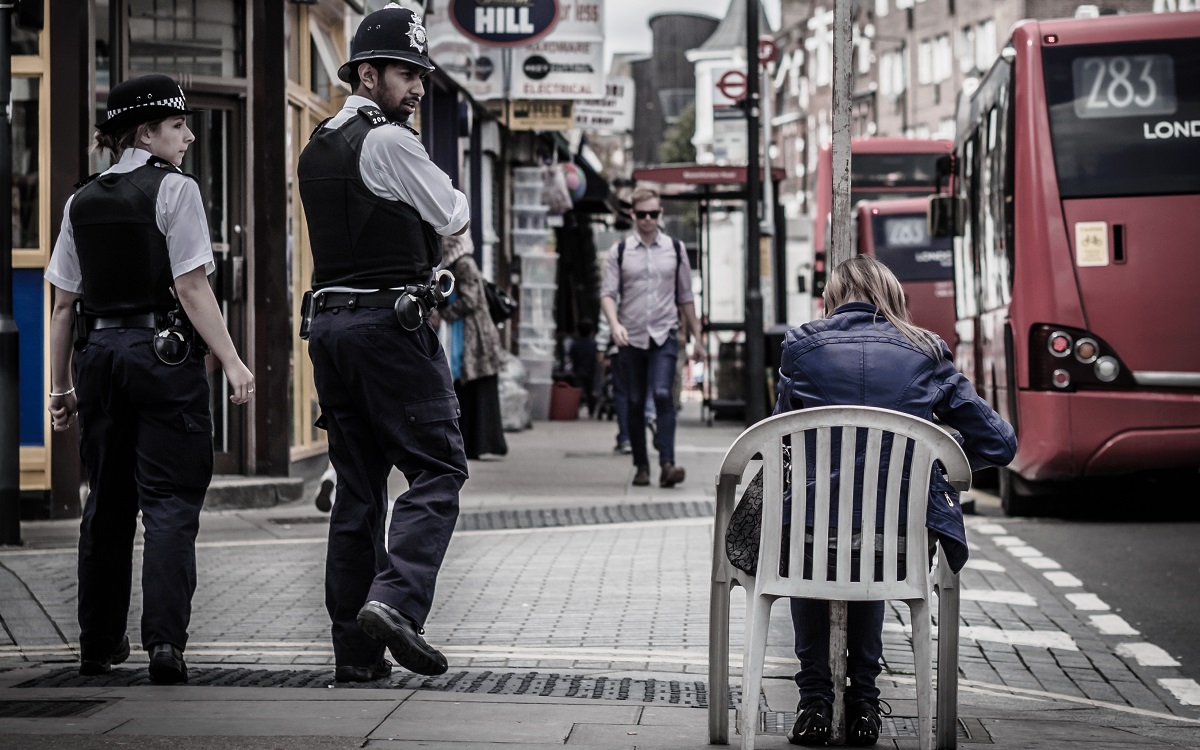 Two police officers looking at a woman sat on a plastic chair on a London street