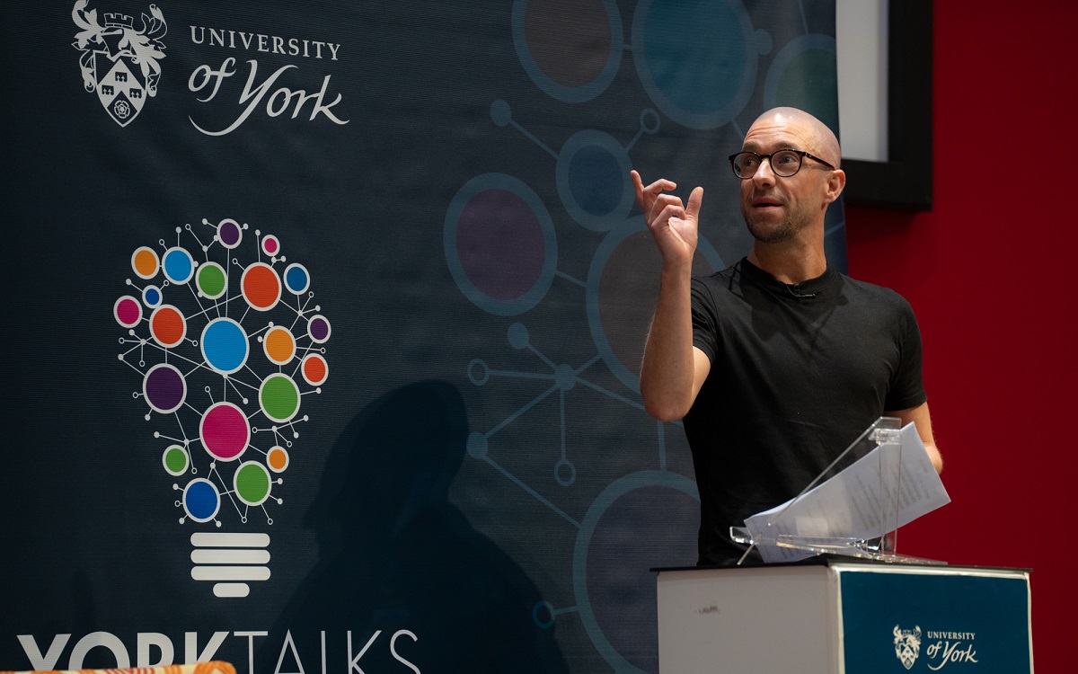 Dr Chris Devany speaking at a lectern with University of York YorkTalks branding in the background