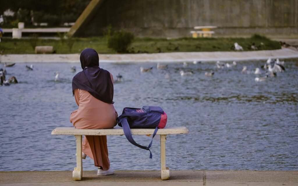 Woman sat on a bench with a backpack staring out at a pond. Photo by kheldoun imad on Unsplash.