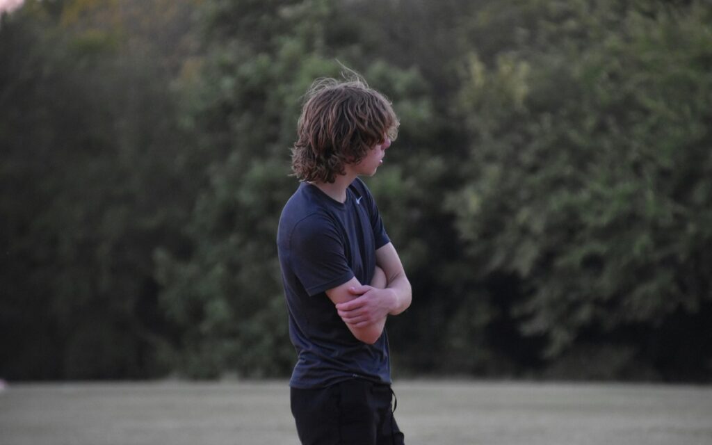 Teenage boy stood with arms crossed alone in a field. Photo by Lesli Whitecotton on Unsplash.
