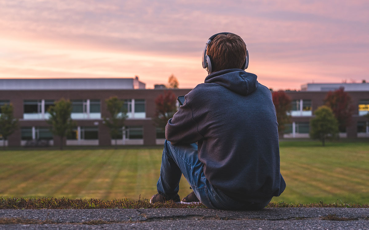 Teenager sat down on ground outside listening to make at sunset.